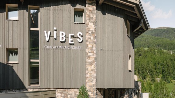 A modern building with a wood and stone facade, surrounded by a green landscape. - VIBES Sölden - your alpine homebase - Sölden