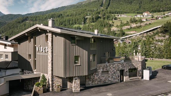 A modern building with the inscription "VIBES" in a picturesque mountain landscape next to a stream. The surroundings are green and the mountains are visible in the background. - VIBES Sölden - your alpine homebase - Sölden