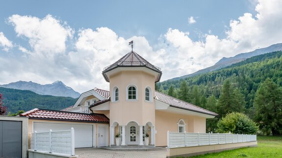 A modern house with a round tower and a red gable roof. In the background, green mountains and a blue sky can be seen. - Villa Castelbell - Sölden
