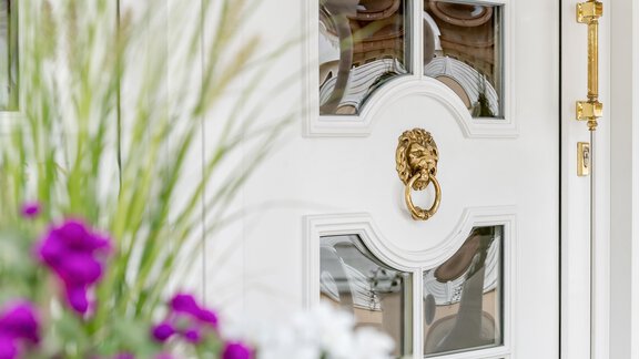 An elegant white door with a golden door knocker in the shape of a lion. Colorful flowers bloom around the door. - Villa Castelbell - Sölden