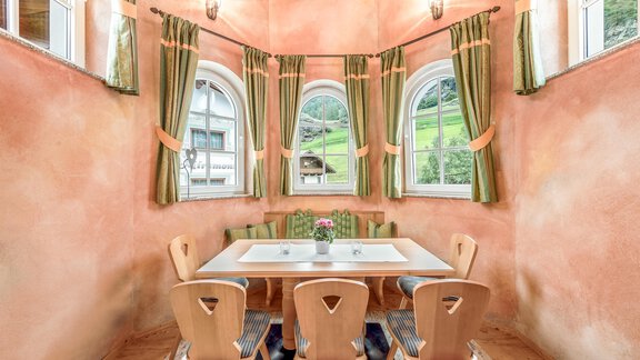 A cozy dining area with a large table and wooden chairs. The walls are painted in warm colors, and large windows offer a view of the greenery. - Villa Castelbell - Sölden
