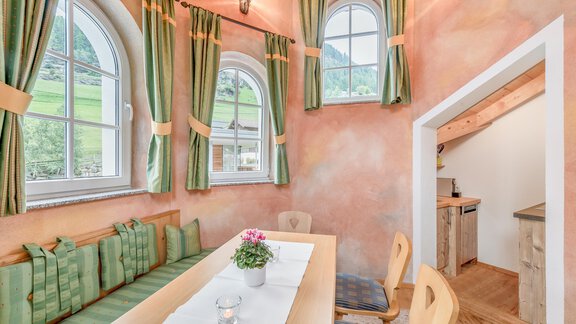 A bright, inviting room with a long table and cozy seating. The walls are painted in soft colors, and there are large windows with green curtains. - Villa Castelbell - Sölden