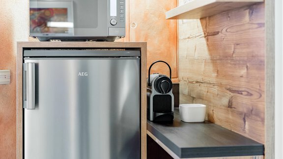 A modern kitchen unit with a refrigerator, a microwave, and a Nespresso machine. The walls are painted in warm colors, and a wooden shelf is visible. - Villa Castelbell - Sölden