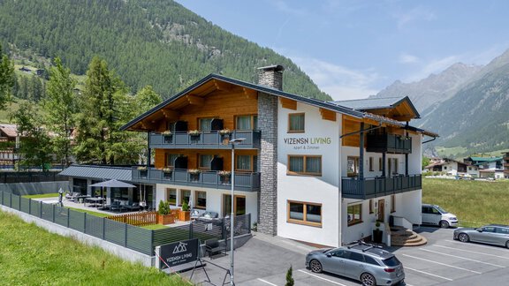 A modern hotel building surrounded by a green meadow and mountains in the background. There is a parking lot with cars and an outdoor seating area. - Vizensn Living - Sölden