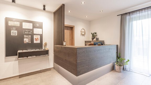A modern reception area with a wooden counter and a board on the wall. Bright lighting and a plant create an inviting atmosphere. - Vizensn Living - Sölden