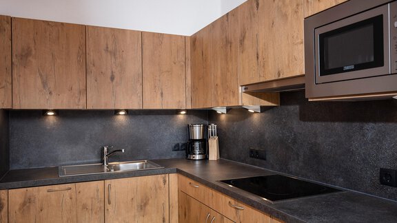 A modern kitchen with wood-colored cabinets and a black countertop. The kitchen is well-lit and equipped with a microwave and a cooktop. - Vizensn Living - Sölden