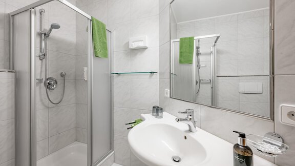 The image shows a modern bathroom with a clear shower and a sink. The walls are brightly tiled, and there are green towels for a fresh accent. - Waldhäusl - Sölden