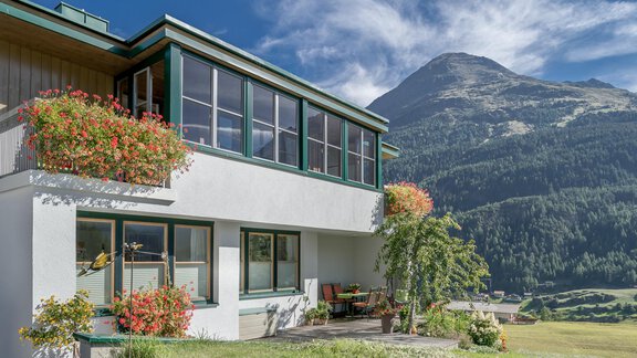 A modern house with large windows and blooming plants. In the background, green mountains stretch under a blue sky. - zuckerhütl NATURAPPART - Sölden