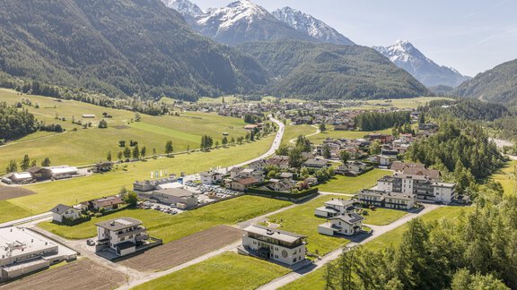 Eine malerische Landschaft mit grünen Feldern und schneebedeckten Bergen im Hintergrund. Im Vordergrund sind moderne Häuser und Straßen zu sehen. - Apart Dorhoam - Umhausen