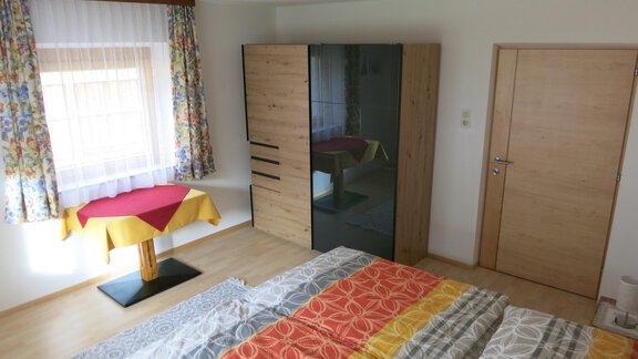 A bright bedroom with a large bed, colorful bedspreads, and a wardrobe. A table with a rotating tablecloth and curtains creates a cozy atmosphere. - Apart Martina - Umhausen