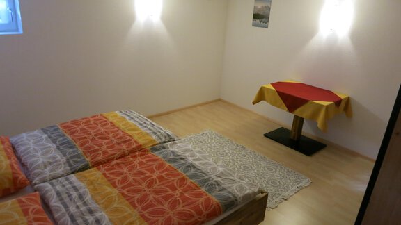 A simple bedroom with a double bed and colorful bedspreads. A table with a red tablecloth stands in the corner. - Apart Martina - Umhausen