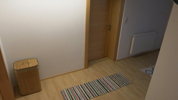 A bright hallway with wooden flooring and a striped doormat. A door and a trash can are also visible. - Apart Martina - Umhausen