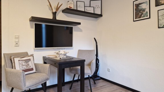 Ein gemütlicher Raum mit einem Fernseher an der Wand und einem kleinen Tisch. Zwei Stühle stehen um den Tisch, und an der Wand hängen Bilder. - Apart Waldnest - Umhausen