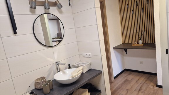 Ein modernes Badezimmer mit einem runden Spiegel und einem stilvollen Waschbecken. Die Wände sind hell gefliest, und es gibt eine Holzverkleidung in einem angrenzenden Bereich. - Apart Waldnest - Umhausen