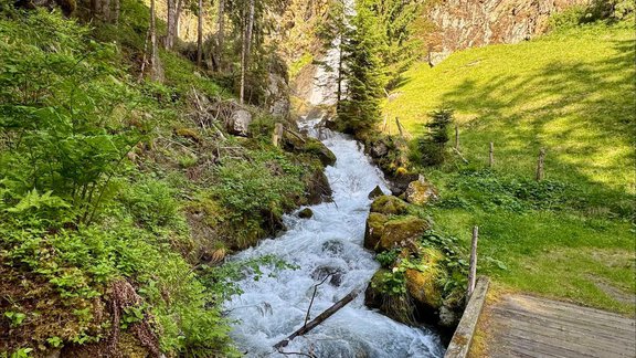 Ein klarer Wasserfluss fließt durch eine grüne Landschaft mit hohen Bäumen. Im Hintergrund sind Felsen und ein kleiner Wasserfall zu sehen. - Appartement Bergglück - Umhausen