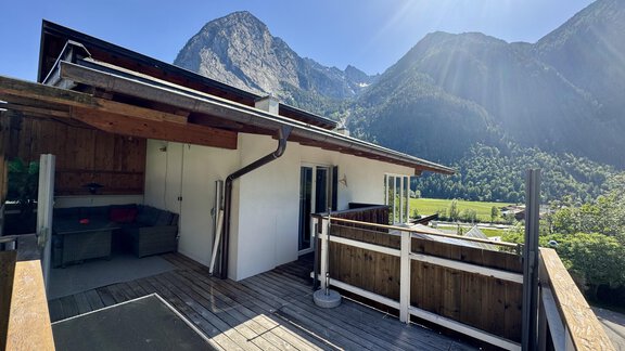 Eine gemütliche Terrasse mit Holzboden und Blick auf hohe Berge. Die Sonne scheint und der Himmel ist klar blau. - Appartement Bergglück - Umhausen