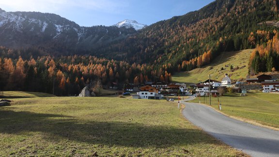 Eine malerische Landschaft mit sanften Hügeln und buntem Herbstlaub. Im Hintergrund sind schneebedeckte Berge und ein kleines Dorf sichtbar. - Appartement Emilia - Umhausen