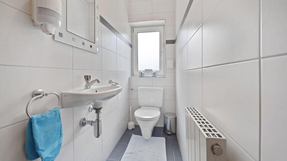 A modern bathroom with white tiles and a bright window. It features a sink, a toilet, and a towel rack with a blue towel. - Biancas Ferienwohnung im Haus Roswitha - Umhausen