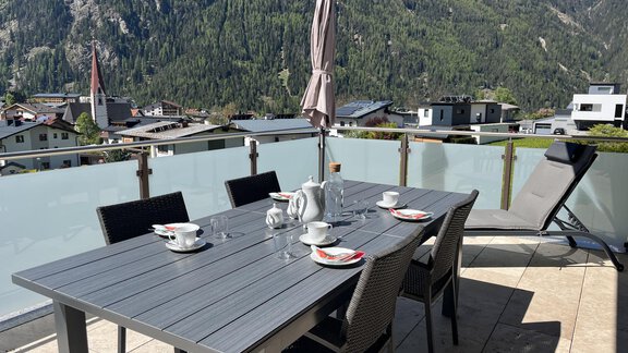 Eine gemütliche Terrasse mit einem großen Tisch, Stühlen und einer Liege. Im Hintergrund sind Berge und eine ruhige Landschaft zu sehen. - BLANKA'S Apart Auszeit - Umhausen