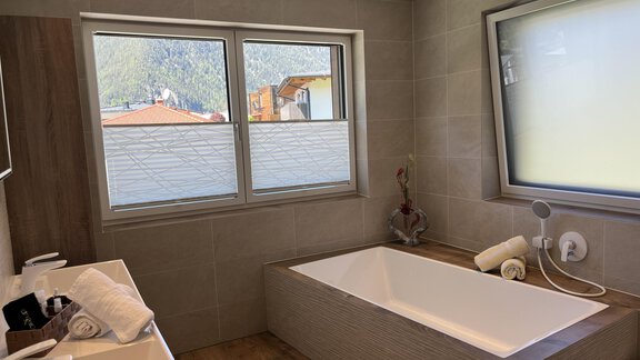 Ein modernes Badezimmer mit einer Badewanne und zwei Fenstern. Die Wände sind hell gefliest und es gibt Handtücher sowie Pflegeprodukte auf der Ablage. - BLANKA'S Apart Auszeit - Umhausen