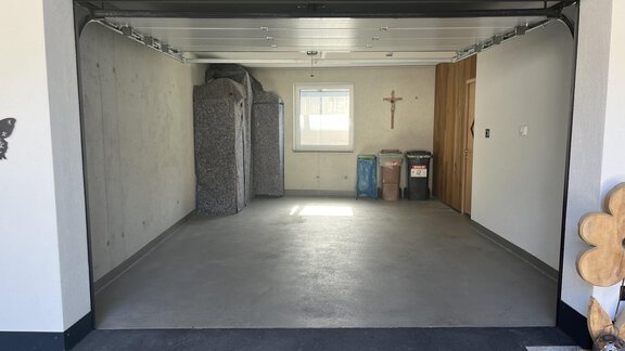 Eine leere Garage mit grauem Boden und Wänden. Auf der linken Seite stehen einige Gegenstände, und an der Wand hängt ein Kreuz. - BLANKA'S Apart Auszeit - Umhausen