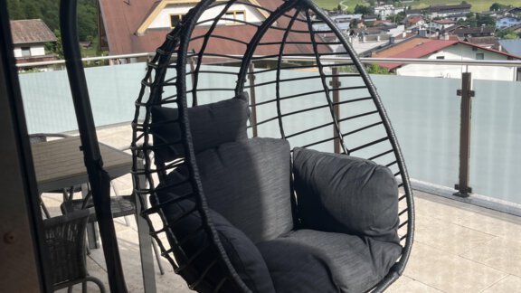 Eine schwingende Hängeschaukel mit schwarzen Kissen steht auf einer Terrasse. Im Hintergrund sind grüne Berge und eine kleine Stadt zu sehen. - BLANKA'S Apart Auszeit - Umhausen