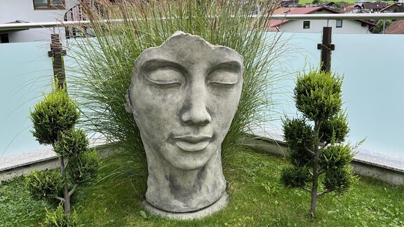A large sculpture of a head stands in a well-kept garden. In the background, trees and a building can be seen, complemented by a blue sky. - BLANKA'S Apart Auszeit - Umhausen