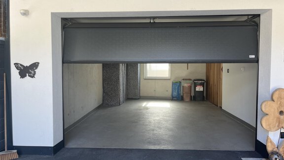 An empty garage with a closed garage door. On the right are some trash cans and a wall with a window. - BLANKA'S Apart Auszeit - Umhausen