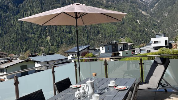 Ein modern eingerichteter Essbereich im Freien mit einem Sonnenschirm. Im Hintergrund sind majestätische Berge und ein blauer Himmel sichtbar. - BLANKA'S Apart Auszeit - Umhausen