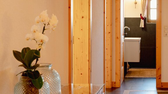 A bright hallway with wooden details and a decorative orchid on a table. In the background, a bathroom is visible. - Chalet Carla - Umhausen