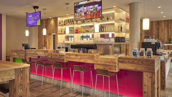 Eine moderne Bar mit Holzdekor und bunten Lichtern. Die Regale sind mit Getränken gefüllt, und Bildschirme zeigen Unterhaltungsinhalte. - Explorer Hotel Ötztal - Umhausen