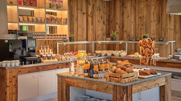 Ein gemütlicher Frühstücksraum mit Holzregalen und einem Buffet. Auf dem Tisch stehen verschiedene Brötchen, Getränke und Snacks bereit. - Explorer Hotel Ötztal - Umhausen