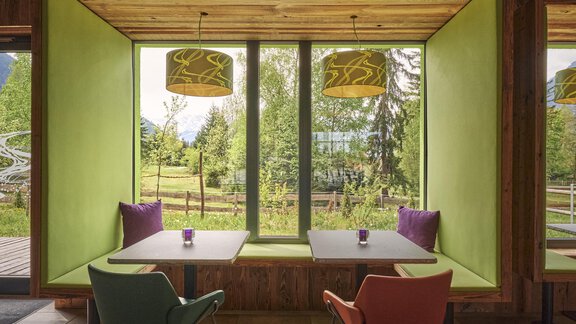 Ein gemütlicher Essbereich mit zwei Stühlen in verschiedenen Farben und einem großen Fenster. Der Raum ist mit grünen Wänden und modernen Lampen dekoriert, und bietet Blick auf die umliegende Natur. - Explorer Hotel Ötztal - Umhausen
