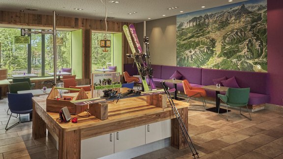 Ein modernes Café mit gemütlichen Sitzgelegenheiten und einer großen Wandkarte der Berge. Die Einrichtung besteht aus Holz und bunten Möbeln, die eine einladende Atmosphäre schaffen. - Explorer Hotel Ötztal - Umhausen