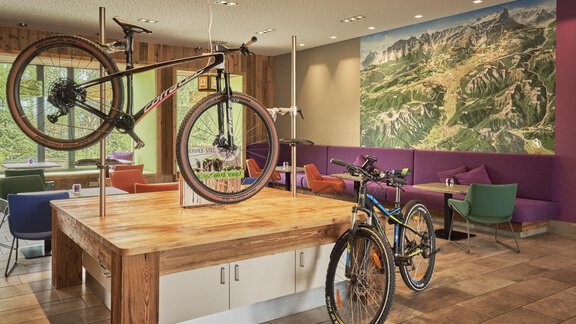 Ein modernes Café mit zwei Fahrrädern, die an einer Holztafel hängen. Im Hintergrund ist ein großes Landschaftsbild zu sehen. - Explorer Hotel Ötztal - Umhausen