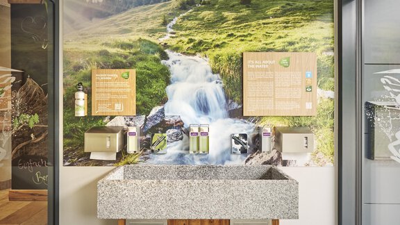 Ein moderner Waschbereich mit einer Steinspüle und einem bergigen Hintergrund. An der Wand sind Informationen und Produkte für die Pflege zu sehen. - Explorer Hotel Ötztal - Umhausen