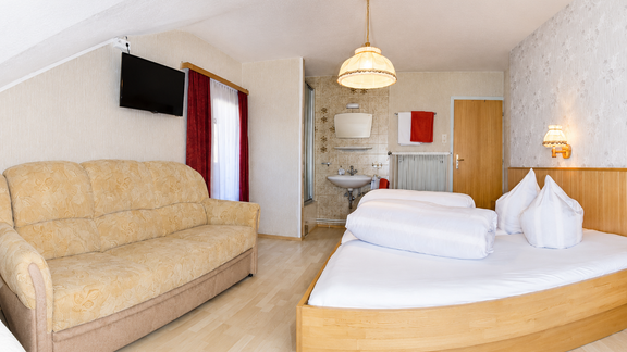 A cozy room with a double bed and a couch. There is a TV on the wall and a bathroom in the background. - Ferienhaus am Spielroan - Umhausen
