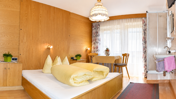 A cozy bedroom with a wood design and a large window. There is a double bed with yellow pillows and a small seating area with a table and chairs. - Ferienhaus am Spielroan - Umhausen