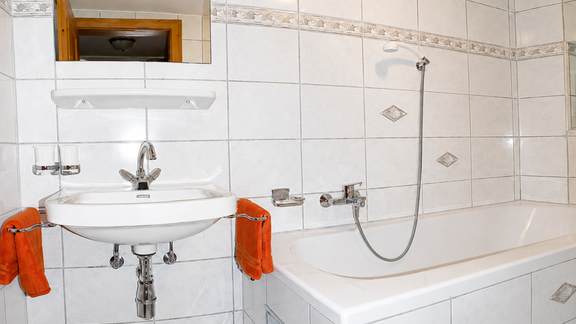 A modern bathroom with white tiles, a bathtub, and a sink. Orange towels hang decoratively next to the sink. - Ferienhaus am Spielroan - Umhausen