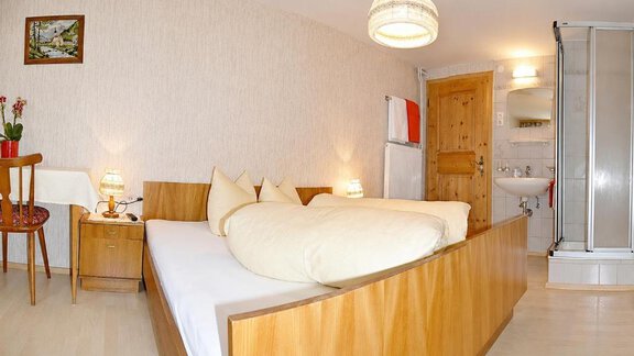 A cozy room with a double bed and wooden details. Next to the bed stands a table with a chair and there is a bathroom with a shower. - Ferienhaus am Spielroan - Umhausen
