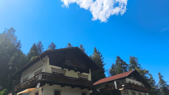 Two traditional houses with wooden-clad balconies under a clear blue sky. Surrounded by green trees and an idyllic mountain landscape. - Ferienwohnung Martha - Umhausen