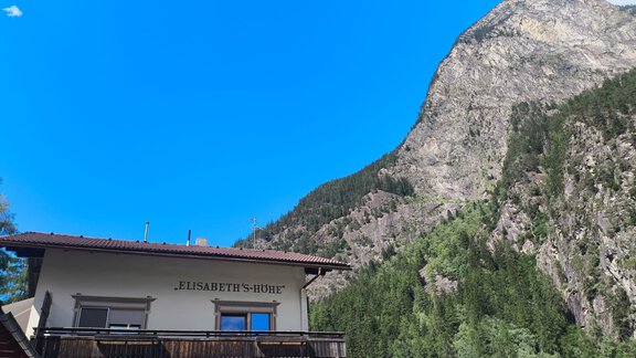 A house with the inscription "Elisabeth's Cabin" stands in front of an impressive mountain landscape. The sky is clear and blue, surrounded by green forests. - Ferienwohnung Martha - Umhausen