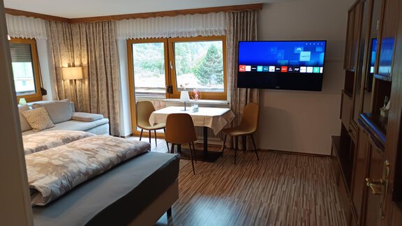 A cozy room with a double bed and a comfortable sofa. On the wall, there is a large television and a table with chairs stands in front of a window. - Ferienwohnung Martha - Umhausen