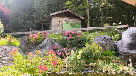 A cozy wooden house in the greenery surrounded by colorful flowers and stones. In the background, tall trees can be seen. - Ferienwohnung Martha - Umhausen
