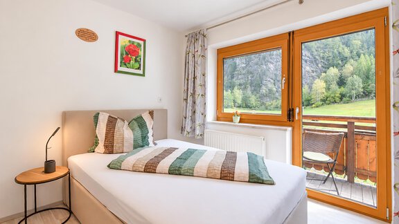 A cozy bedroom with a single bed and colorful pillows. Large windows offer a view of the green landscape outside. - Ferienwohnung Martha - Umhausen