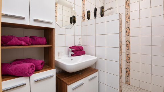 A modern bathroom with white tiles and a sink.  
Pink towels are neatly placed on a shelf. - Ferienwohnung Martha - Umhausen