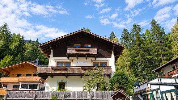 A traditional house in the mountains with balconies and surrounded by trees. The sky is clear and blue, which emphasizes the tranquil atmosphere. - Ferienwohnung Martha - Umhausen
