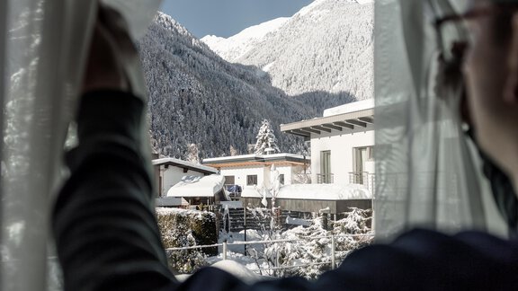Eine Person öffnet den Vorhang und blickt auf eine verschneite Berglandschaft. Im Vordergrund sind Häuser und die schneebedeckten Berge im Hintergrund zu sehen. - MYALPS Ötztal - Umhausen