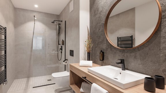 Ein modernes Badezimmer mit einer begehbaren Dusche und einem eleganten Waschbecken. Die Wände sind in neutralen Grautönen gehalten, und ein runder Spiegel ergänzt das Design. - MYALPS Ötztal - Umhausen