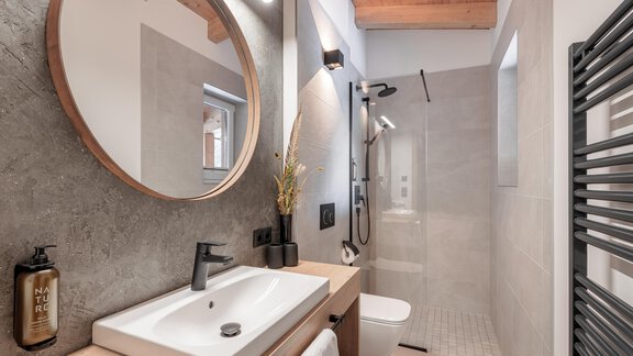 Ein modernes Badezimmer mit einem runden Spiegel und einer stilvollen Dusche. Die Wände sind in neutralen Tönen gehalten und es gibt Holzdetails an der Decke. - MYALPS Ötztal - Umhausen